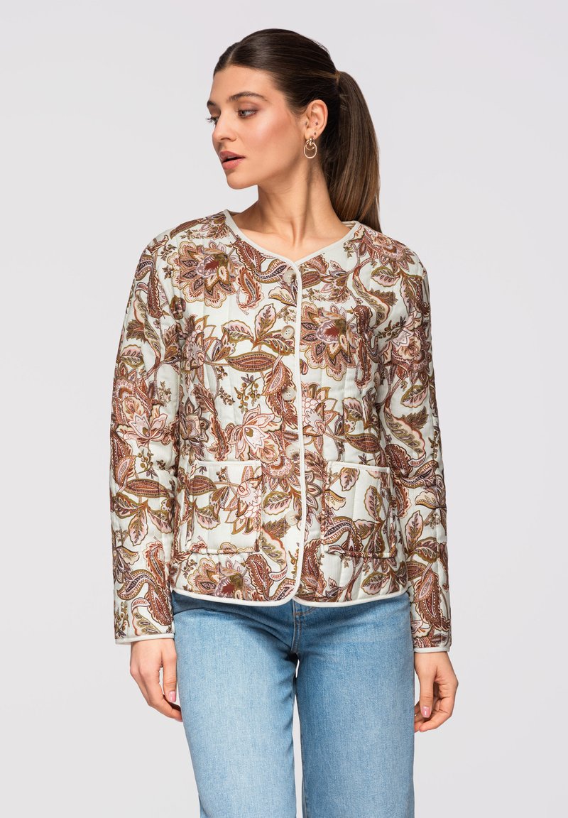Femme portant une veste boutonnée avec des motifs floraux marron et rose, associée à un jean bleu clair, regardant vers sa gauche.