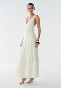 Witte maxi-jurk met een V-hals, gestructureerde stof en een gelaagde zoom. Gecombineerd met doorzichtige sandalen met bandjes en dikke armbanden.