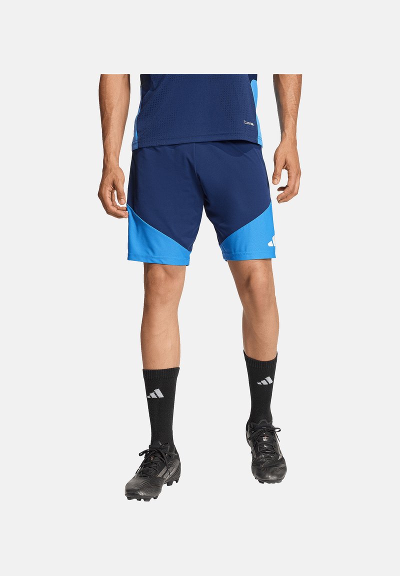 Homme portant un short de sport bleu marine et bleu clair, des chaussettes Adidas noires et des crampons noirs, debout devant un fond uni.