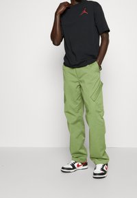 Zwarte T-shirt met een rood Jordan logo, gecombineerd met lichtgroene cargobroek. Kenmerken zijn onder andere zakken en donkere sneakers met witte en rode accenten.