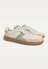 Light beige sneakers with a textured surface, green accents, and a brown rubber sole. Features lace-up design and padded collar.