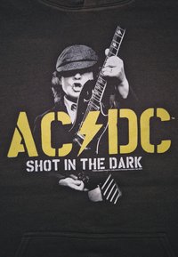 Muž v kšiltovce hraje na elektrickou kytaru s odvážným žlutým logem AC/DC a bílým textem "Shot in the Dark" na černém pozadí.