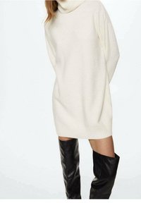 Overmaat crème coltrui met geribde zoom, gemaakt van zacht textuurmateriaal, gecombineerd met glanzende zwarte laarzen tot boven de knie.