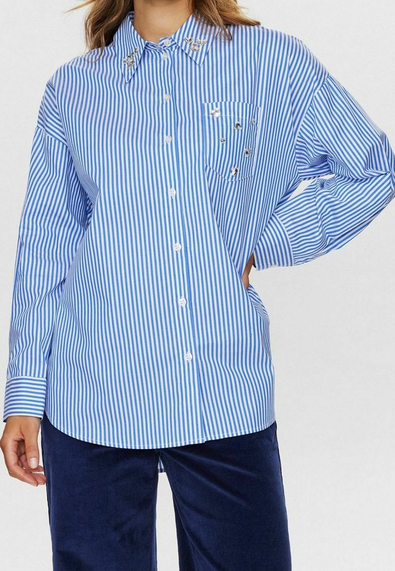 Femme portant une chemise à rayures verticales bleues et blanches boutonnée, avec des détails en strass sur le col et la poche poitrine gauche, associée à un pantalon bleu foncé.