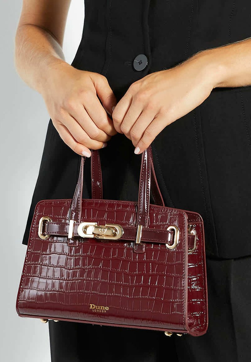 Handen die een bordeauxrood lederen handtas met krokodillenembossing en gouden hardware vasthouden, gedragen met een zwarte knoopvest.