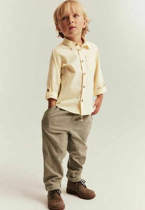 Jeune garçon aux cheveux blonds portant une chemise jaune à boutons retroussée, un pantalon beige et des bottes marron à lacets, debout les mains dans les poches.