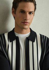 Jachetă din tricot cu dungi verticale alb-negru, guler ascuțit și fermoar frontal; purtată peste un tricou alb simplu.