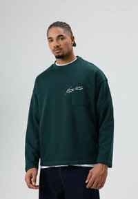 Grüner Pullover mit Rundhalsausschnitt, langen Ärmeln und einer vorderen Tasche. Auf der Brust befindet sich ein weißes Logo in Schriftzug. Glatte Textur.