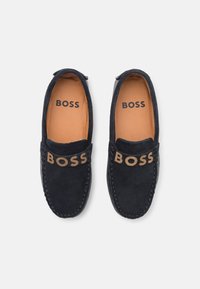 Tumši zila zamšāda loafers ar brūnām iekšējām zolēm, liels "BOSS" logo brūnā krāsā priekšējā siksniņā un melns "BOSS" teksts iekšpusē.