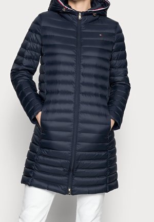 Kobieta w granatowej pikowanej długiej kurtce puchowej z kapturem, z rękami w kieszeniach, w białych spodniach.