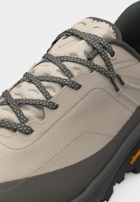 Ελαφρώς μπεζ παπούτσι πεζοπορίας με σκούρα γκρι σόλα και γκρι κορδόνια με σχέδια, δεμένα πάνω από τη μαλακή υφή της γλώσσας του παπουτσιού.