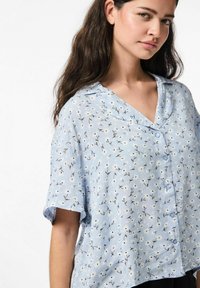 Mėlyna trumpomis rankovėmis bluzonė su V formos iškirpte, papuošta gėlių raštu ir mygtukų užsegimu priekyje. Lengvas audinys su laisvu siluetu.