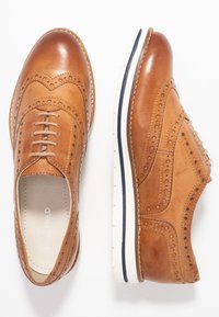 Zapatos oxford de cuero marrón con detalles de brogue, punta redonda, suela blanca plana y cordones marrones. Textura suave con acentos de costura visibles.