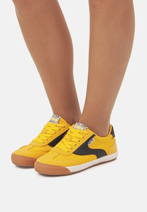 Zapatillas de lona amarillas con un panel lateral negro, suela de goma, puntera redonda y detalles blancos en la suela, con un diseño de cordones.