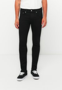Hombre vistiendo jeans negros ajustados y zapatillas negras con cordones blancos, de pie frente a un fondo blanco liso.