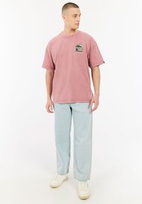 Roze katoenen T-shirt met korte mouwen en een borstzak met een camouflagepatroon. Gecombineerd met lichtblauwe, relaxed-fit jeans en witte sneakers.