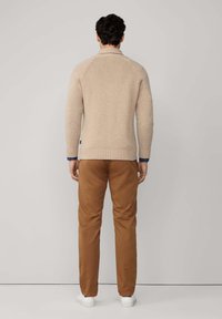 Man staat met zijn rug naar de camera, gekleed in een beige gebreide trui, bruine broek en witte sneakers, tegen een effen lichtgrijze achtergrond.