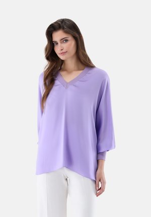 Frau mit langen braunen Haaren, die eine lockere, hellviolette Bluse mit V-Ausschnitt und weiße Hose vor einem einfarbigen Hintergrund trägt.