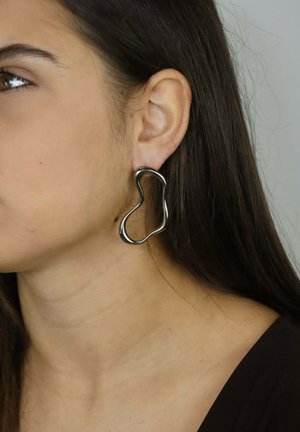 Abstract gevormde zilveren oorbellen met een gladde, glanzende textuur, voorzien van een doorlopende lus. Draagbaar aan het linkeroor van een model.