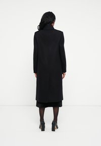 Lungo cappotto nero con colletto strutturato, design sartoriale e tessuto liscio. Presenta una cucitura centrale sul retro e maniche aderenti.