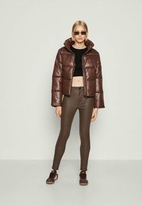 Young woman wearing brown shiny puffer jacket, black cropped top, brown leather pants, purple sneakers, and rectangular sunglasses, standing straight.
