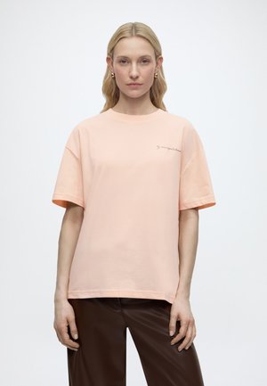 Blondynka w dużym brzoskwiniowym t-shircie z małym napisem na piersi i ciemnobrązowych spodniach, stojąca na jasnym, jednolitym tle.