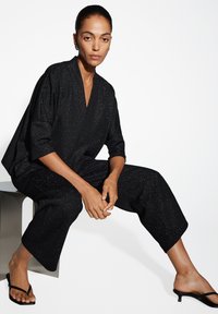 Zwarte, glanzende stoffen outfit met een losse pasvorm, diepe V-hals en wijde mouwen. Gecombineerd met platte, gescheurde sandaal.