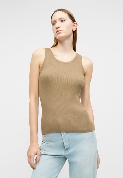 Νεαρή γυναίκα με ίσια ξανθά μαλλιά που φοράει μπεζ ριγωτό τοπ χωρίς μανίκια και ανοιχτόχρωμο μπλε τζιν, στέκεται μπροστά σε λευκό, μονόχρωμο φόντο.