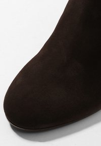 Bottine en daim marron foncé avec bout arrondi sur fond clair.