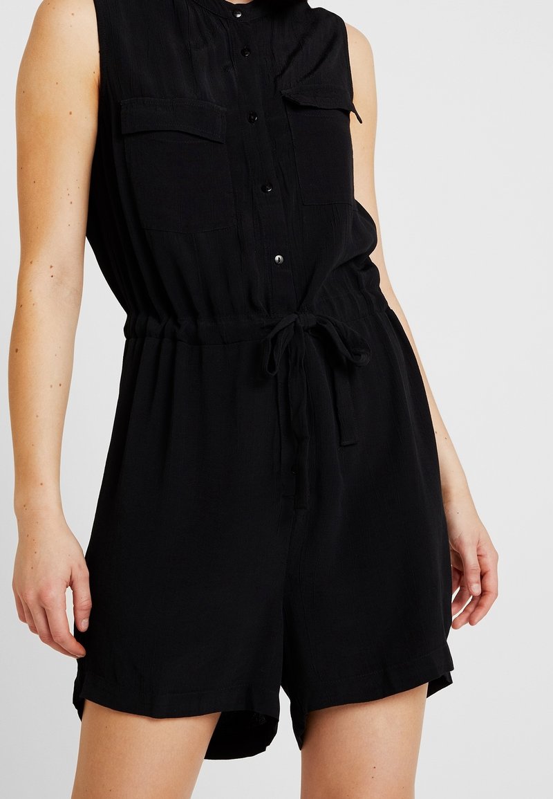 Combinaison noire sans manches à boutons sur le devant, avec poches poitrine et ceinture nouée à la taille, portée par une personne sur un fond clair uni.