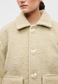 Beige, textúrált kabát, galléros nyakkal, szív alakú gombokkal és zsebekkel. Puha, bolyhos anyaga melegséget biztosít.