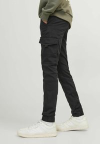 Pantalons cargo noirs avec des poignets élastiques, poches latérales et une coupe décontractée. Fabriqués en tissu durable. Portés avec des baskets blanches.