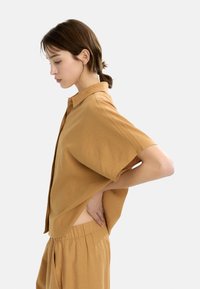 Kobieta w luźnej brązowej koszuli i spodniach stoi w profilu z ręką na dolnej części pleców na tle jednolitego białego tła.