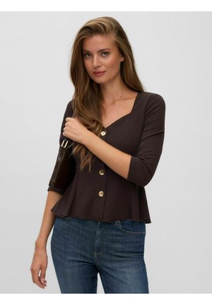 Giovane donna con lunghi capelli castano chiaro indossa un top marrone scuro con bottoni e jeans blu, tenendo una borsa scura sulla spalla.