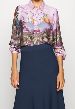 Hemd mit Knöpfen, das ein Blumen- und Landschaftsmuster in Rosa- und Grüntönen zeigt. Lange Ärmel mit gerafften Manschetten, kombiniert mit einem marineblauen Rock.