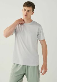 Light gray short-sleeve athletic shirt with a textured pattern, round neckline, and small black logo at the hem. Worn with gray shorts.