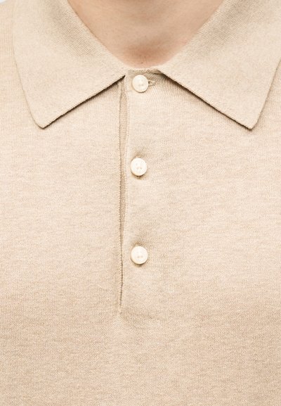 Tricou polo bej din tricot cu trei nasturi crem și guler ascuțit, purtat pe partea superioară a pieptului și gâtului unei persoane.