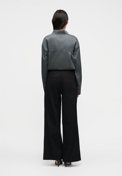 Γυναίκα με μακριά σκούρα μαλλιά που φοράει γκρι μακρυμάνικη μπλούζα μέσα σε μαύρο παντελόνι ψηλόμεσο με φαρδιά μπατζάκια και μαύρες γόβες, στεκόμενη με την πλάτη προς τα εμπρός.