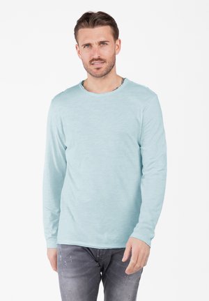 A man with short hair wears a light blue long-sleeve shirt and gray jeans, standing against a white background, looking forward.