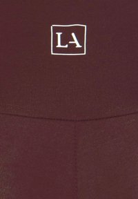 Burgundy activewear fabric featuring a white square logo "LA" on the waistband, with a smooth texture and stitched seam detail.
