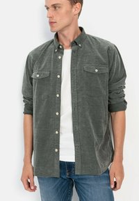Graues Cordhemd mit Button-Down-Kragen, zwei Brusttaschen und aufgerollten Ärmeln. Kombiniert mit einem weißen T-Shirt und blauen Jeans.