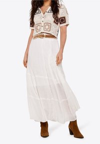 Žena na sobě má bílou maxi sukni se sklady, háčkovaný top s hnědými vzory, světlý opasek a hnědé semišové kotníkové boty.