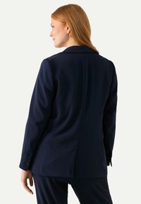 Blazer navy con una vestibilità su misura, spalle strutturate e polsini con tre bottoni. Tessuto liscio, spacchetto singolo sul retro, design minimalista.