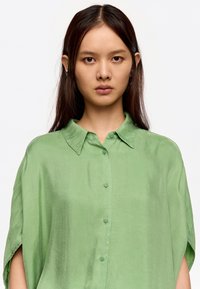 Camisa verde de botones de manga corta, hecha de una tela suave con cuello y un ligero brillo. Presenta un corte holgado y aberturas en los laterales.