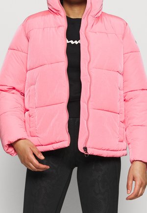 Persona que lleva una chaqueta acolchada rosa brillante sin cerrar, sobre una camiseta negra y leggings negros con estampado, de pie frente a un fondo liso.