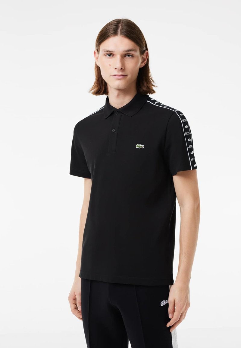 Camisa polo negra de algodón, con cuello clásico, mangas cortas y franjas de acento blancas en los hombros. Logo verde en el pecho.