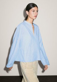 Une femme marche avec confiance, portant un haut bleu clair avec un col mandarin et un pantalon beige, sur un fond neutre simple.