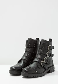 Černé kožené kotníkové boty se třemi nastavitelnými pruhy a stříbrnými přezkami, zdobené hřebíky na špičce a po stranách, s texturovaným povrchem.