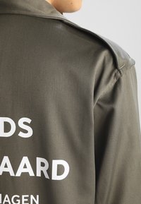 Veste vert olive au toucher lisse, dotée d'un large col et d'une inscription blanche dans le dos. Le matériau semble léger et durable.