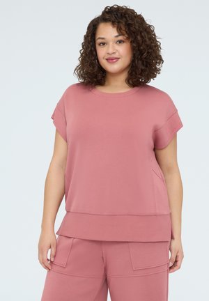 Frau mit lockigem Haar, die ein lockeres, kurzärmeliges rosa Top und passende rosa Cargo-Shorts trägt, steht vor einem hellen Hintergrund.